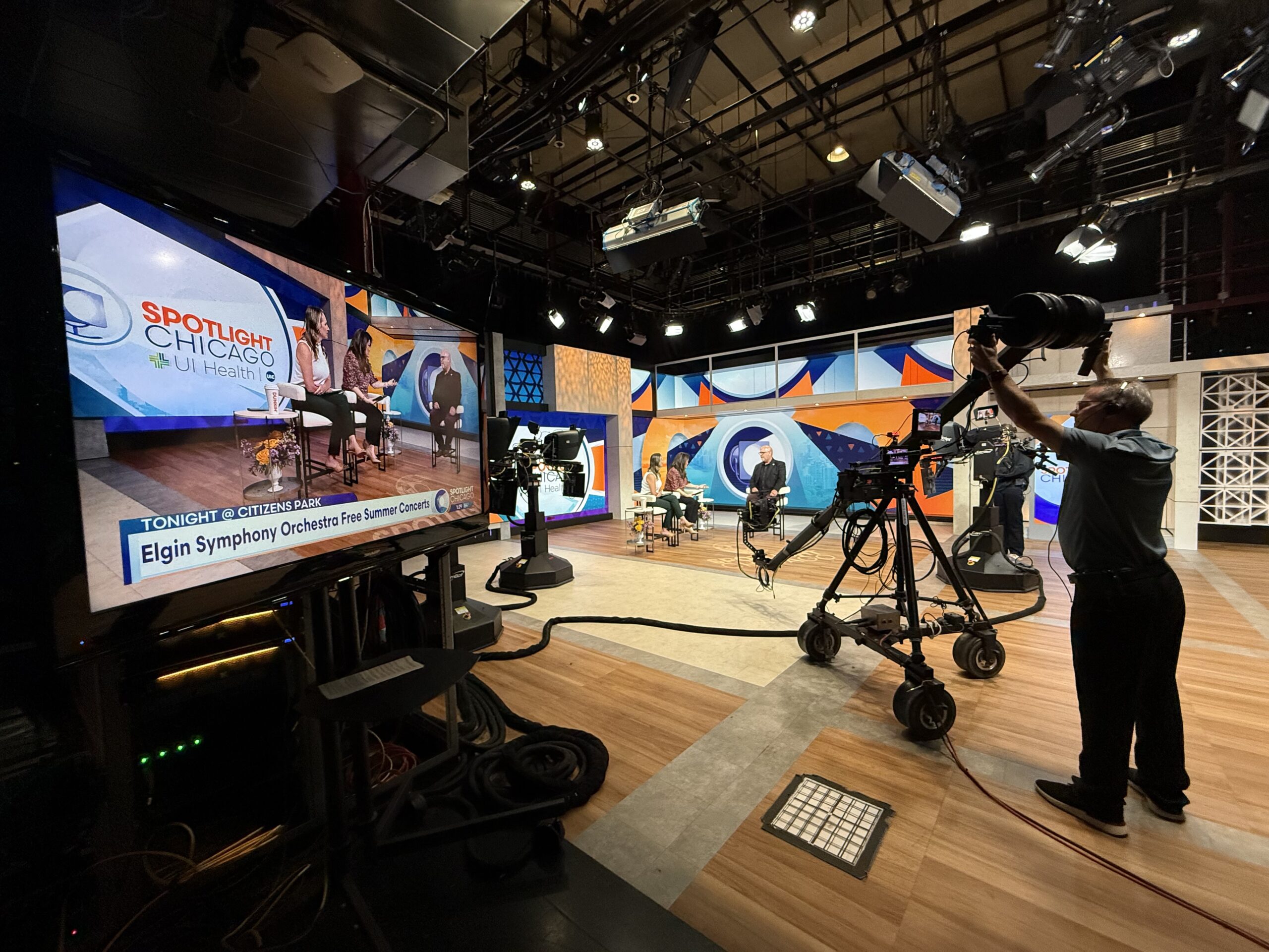 A photo of a TV studio, with a TV screen identifying the Elgin Symphony Orchestra on the left of the screen, along with 2 female anchors and one male who was being interviewed, and a large TV camera on a dolly, being operated by a WGN-TV camera operator.